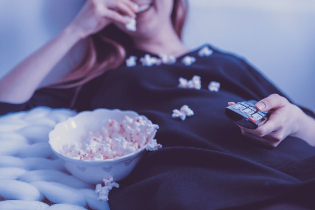 Woman lying back with remote in left hand, eating popcorn with right hand.